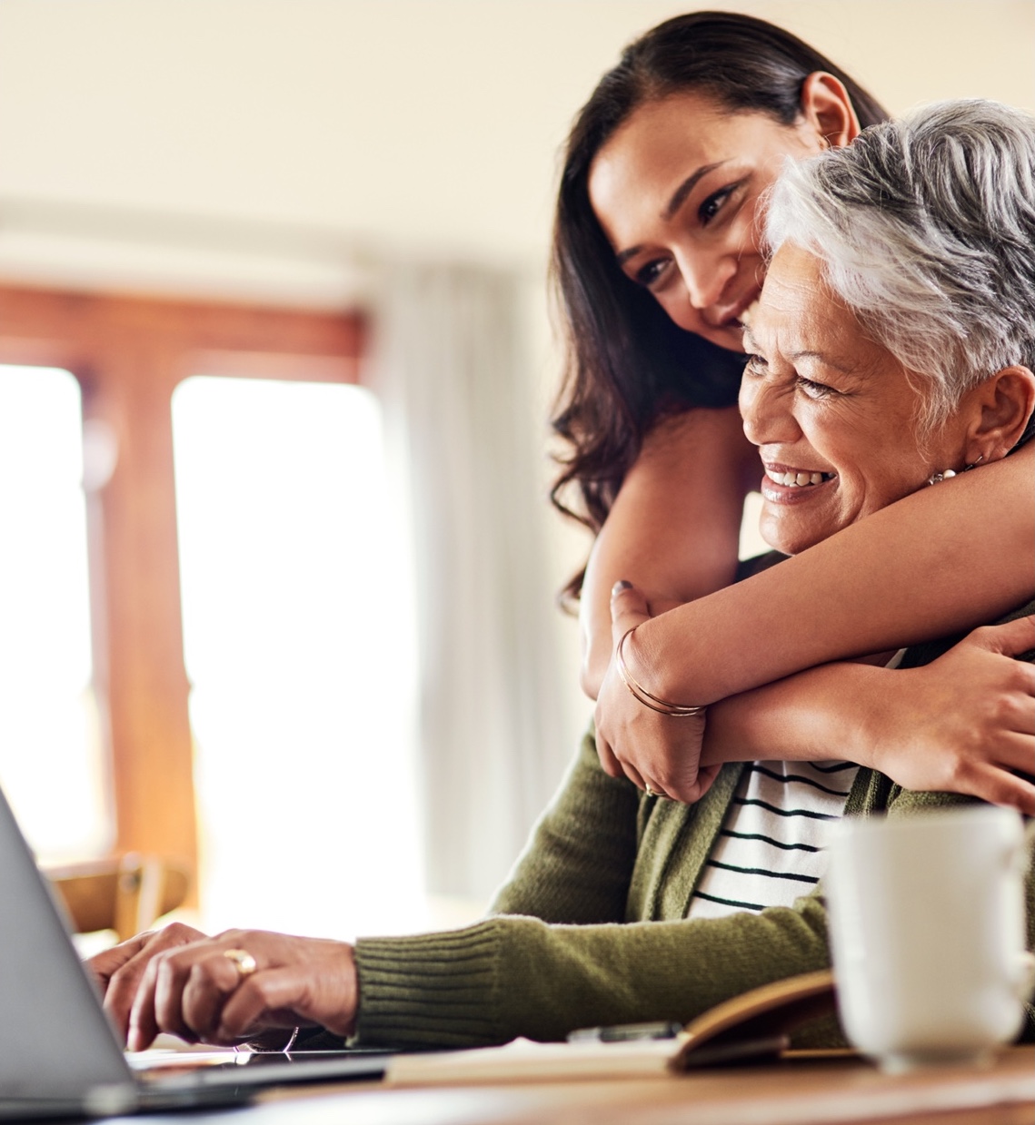 a young woman hugs her mother while she uses her laptop